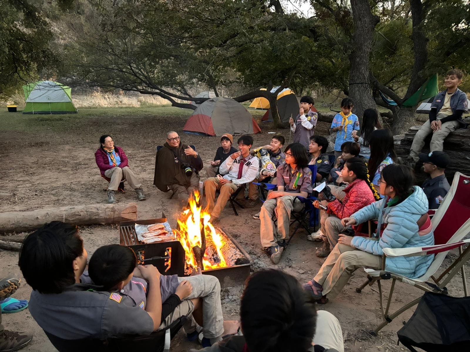 Scouts and trưởng around a campfire at dusk, tents pitched among the trees