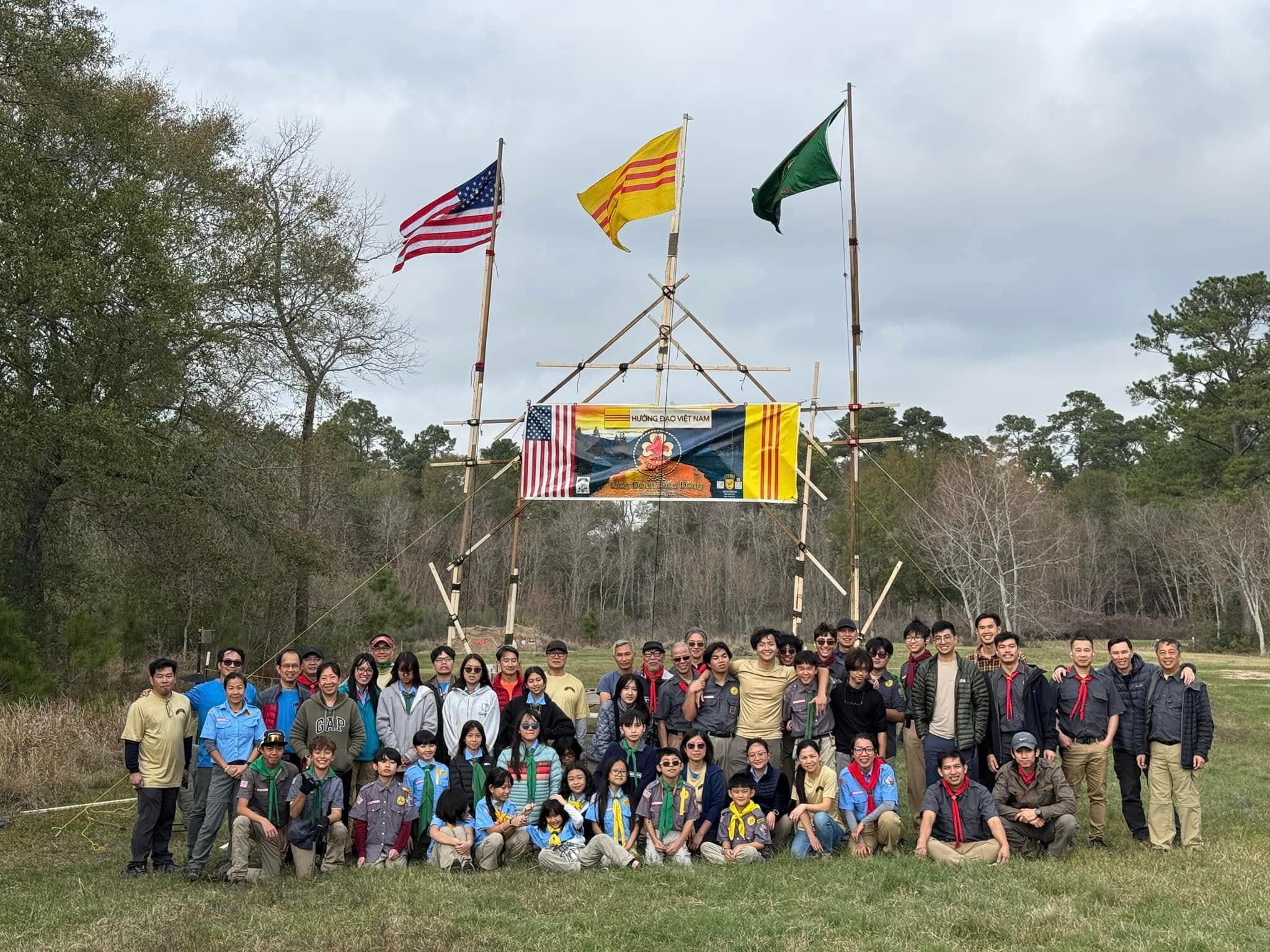 Scouts and families gathered beneath the American flag, the Vietnamese heritage flag, and the scouting flag at a lashed-timber camp gateway